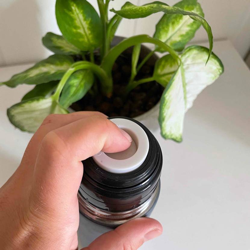 Thermosflasche im Test: Eine Hand drückt auf die Öffnung einer dunkelblauen Thermosflasche, welche vor einer Zimmerpflanze auf einem weißen Tisch. steht.