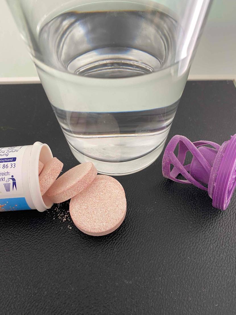 Multivitamin-Tabletten im Test: Auf einer dunklen Oberfläche steht ein Glas, daneben eine geöffnete Packung Multivitamin-Tabletten und der Deckel