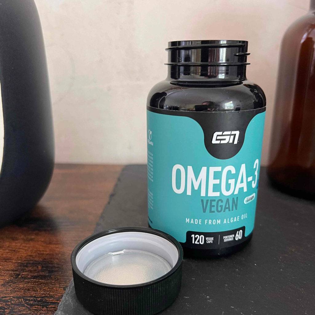 Omega-3-Kapseln im Test: Dunkle Dose mit hellbaluen Logo und geöffnetem Deckel steht auf einem Schreibtisch.