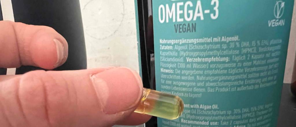 Omega-3-Kapseln-Test: Hand hält eine gelb-durchsichtige Omega-3-Kapsel vor die hellblaue Omega-3-Kapsel-Dose.