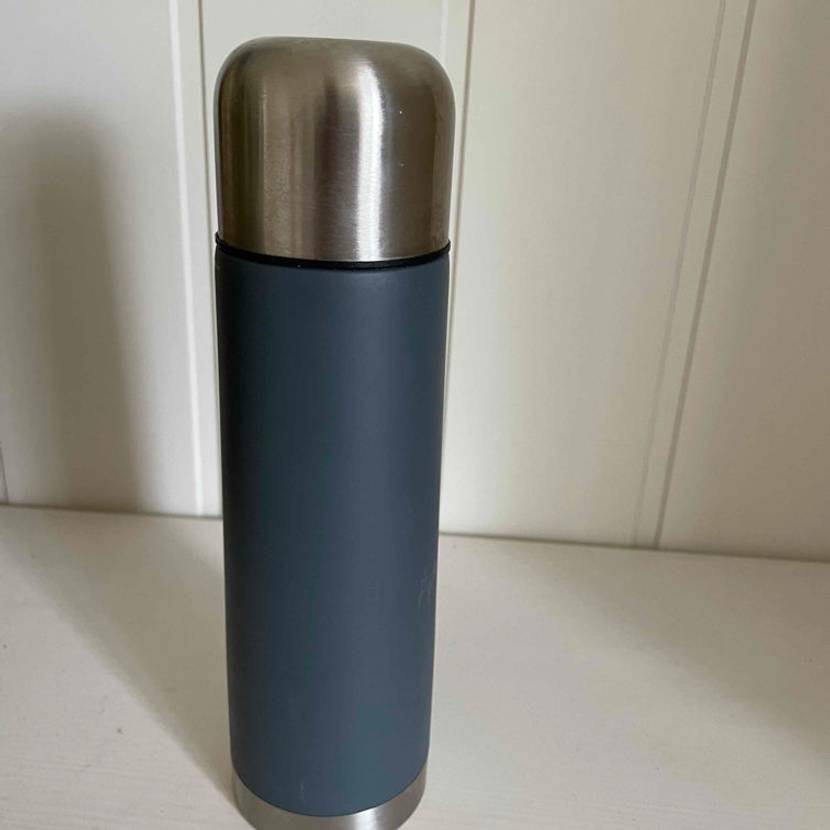 Getestete Thermosflasche in dunkelblau mit Edelstahldeckel steht vor einer weißen Wand.
