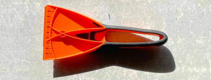 Rückwärtige Ansicht eines orangen Eiskratzers im Test inklusive schwarz gummiertem Handgriff, der auf melierter Oberfläche liegt.