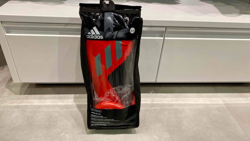 Schwarz-rote Adidas Boxhandschuhe im Test lehnen in ihrer schwarzen Kunststoffverpackung inklusive Sichtfenster am Rand eines weißen Verkaufsregals.