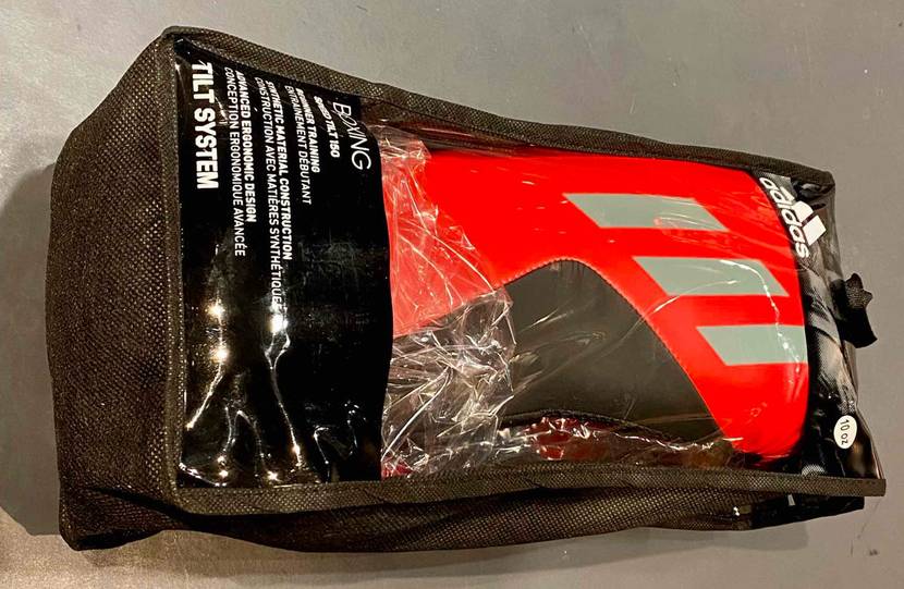 Adidas Boxhandschuhe im Test: ein schwarz-rotes Modell mit silbernen Streifen liegt innerhalb seiner schwarzen Verpackung inklusive Sichtfenster auf einem grauen Untergrund.