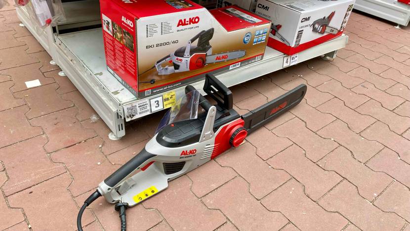 Eine Al-Ko Elektro-Kettensäge im Test in rot-grauen Design inklusive schwarzer Schwertschutz-Hülle steht vor einem Verkaufsregal mit der Originalverpackung auf roten Steinfliesen.