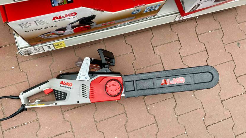 Ansicht von oben auf eine auf roten Steinfliesen liegende Al-Ko Elektro-Kettensäge in Rot-Grau mit schwarzer Schwertschutz-Hülle.