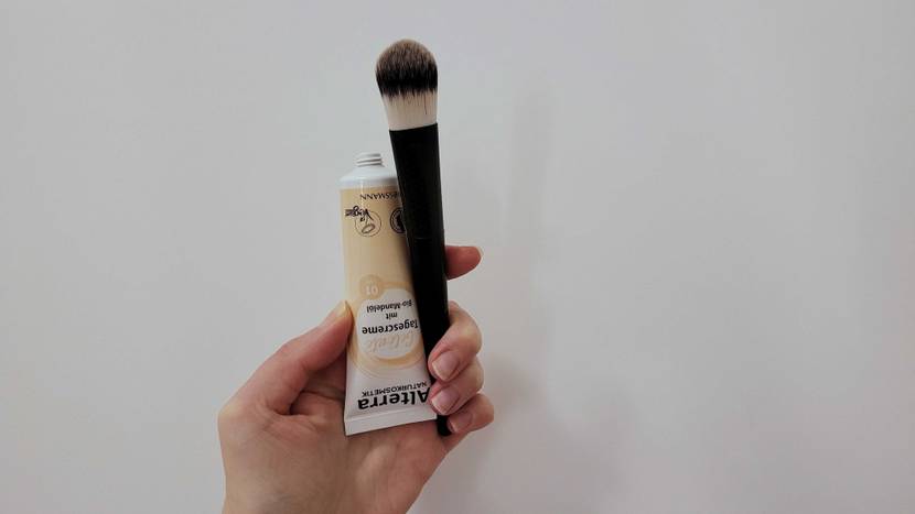 Im Test: Eine Hand hält einen Pinsel und eine Tube getönte Tagescreme vor eine helle Wand.