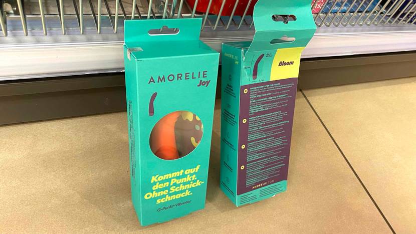 Vibrator-Test: zwei Amorelie Vibratoren stehen nebeneinander in ihren hellgrünen Verpackungen auf einem beigefarben Boden vor einem Verkaufsregal.