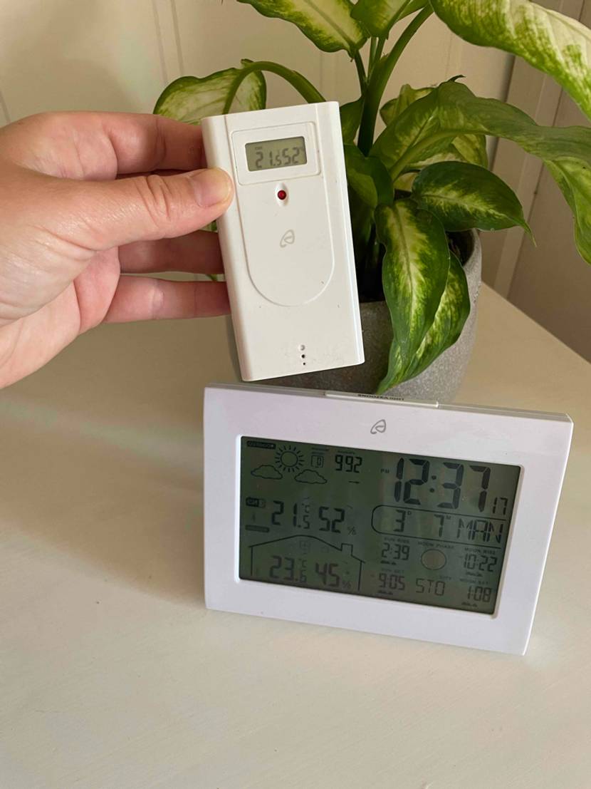 Wetterstation im Test: Hand hält eine Fernebdienung vor eine Wetterstaio, die vor einem Blumentopf auf einem weißen Tisch steht.