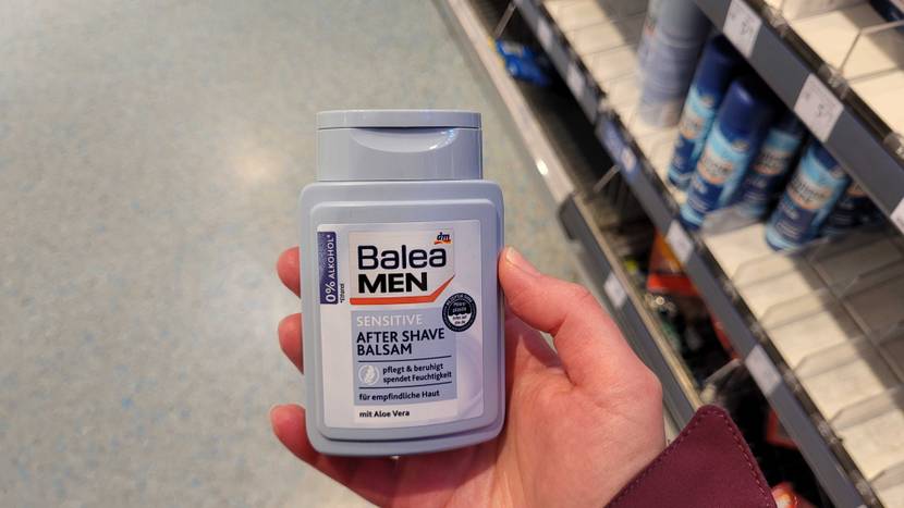 Hand zeigt eine hellblaue Kunststoffflasche, auf der ein Etikett eines Balea Men Aftershaves im Test klebt, vor einem Verkaufsregal.