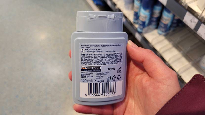Hand hält eine hellblaue Kunststoffflasche, mit der rückwärtigen Produktbeschreibung von Balea Men Aftershave im Test vor einem Verkaufsregal in die Kamera.