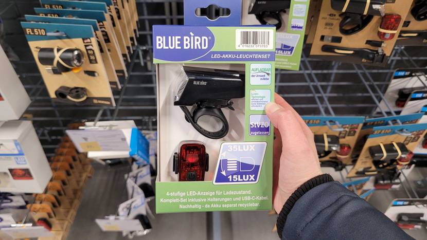 Fahrradlampen-Test: Eine Hand hält eine hellgrün-blaue Fahrradlampen-Verpackung der Marke Blue Bird mit einem LED-AKKU-Leuchtenset vor einem Regal mit verschiedenen Fahrradlampen.