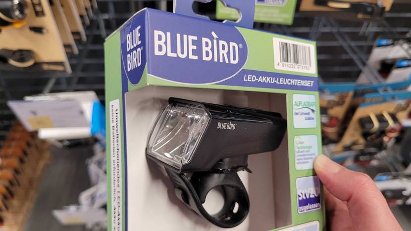 Hand hält ein getestetes Fahrradlampen-Set der Marke Blue Bird, mit Fokus auf die Frontlampe in einer hellgrün-blauen Verpackung vor ein Verkaufsregal.