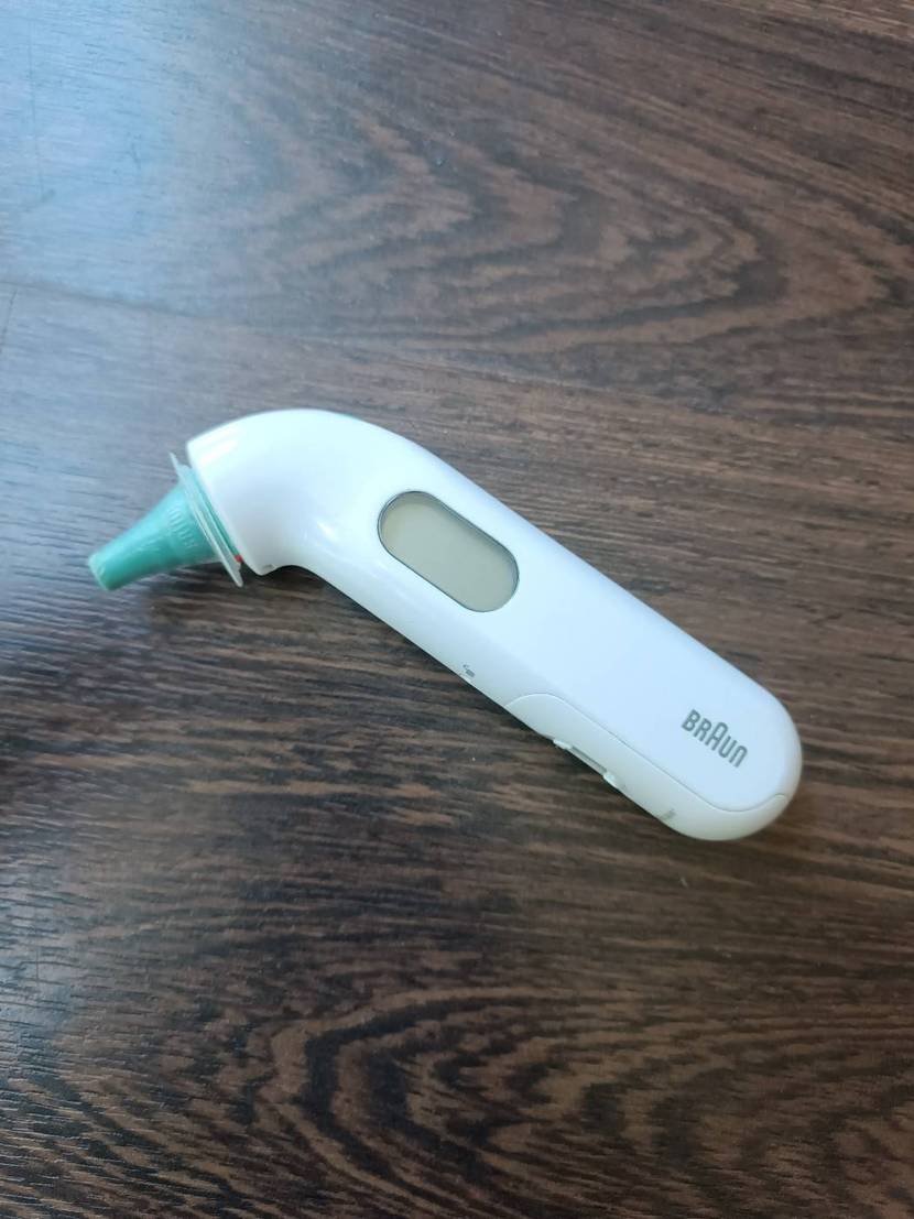 Fieberthermometer-Test: weißes Braun Fieberthermometer mit LCD-Anzeige und grünem Endstück für die Messung im Ohr liegt auf einem dunklen Holzuntergrund.