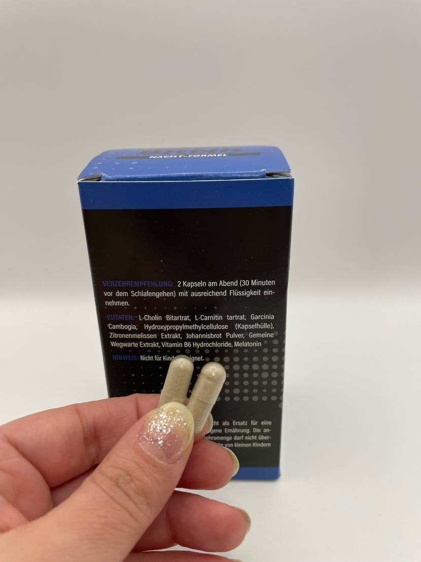 Fatburner-Test: Eine Hand hält zwei Kapseln vor einer mit der Rückseite nach vorn stehenden Verpackung.