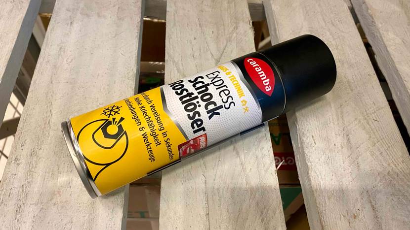 Ein Rostlöser im Test der Marke Caramba liegt in einer Sprühdose, mit seitlich befestigter Sprühverlängerung auf Holzbrettern.