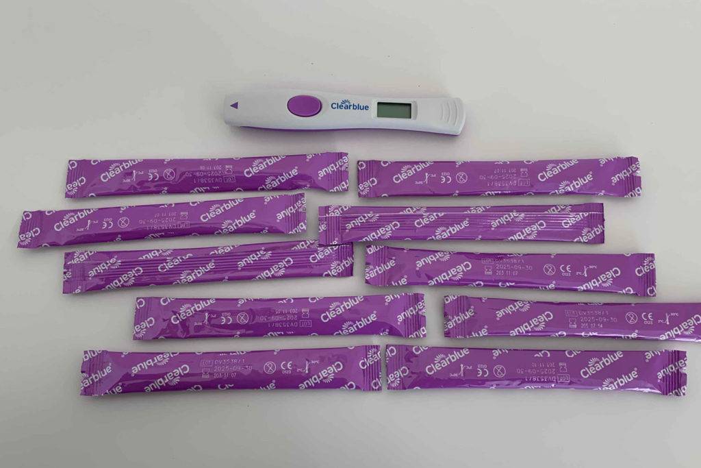 Ein ausgepackter Ovulationstest von Clearblue liegt auf einem weißen Untergrund, darunter in 2er Reihen 10 lila verpackte Tests.