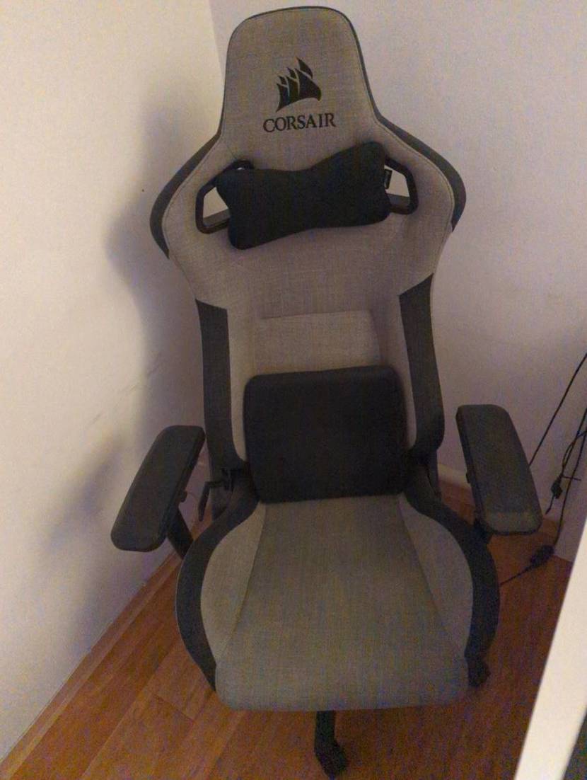 Vorderansicht eines hellgrauen, mit dunklen Elementen abgesetzten Bezugs eines Corsair T3 Rush Bürostuhl im Test inklusive dunkelgrauer Kissen, der auf einem Holzboden zwischen weißer Wand und Tisch steht.