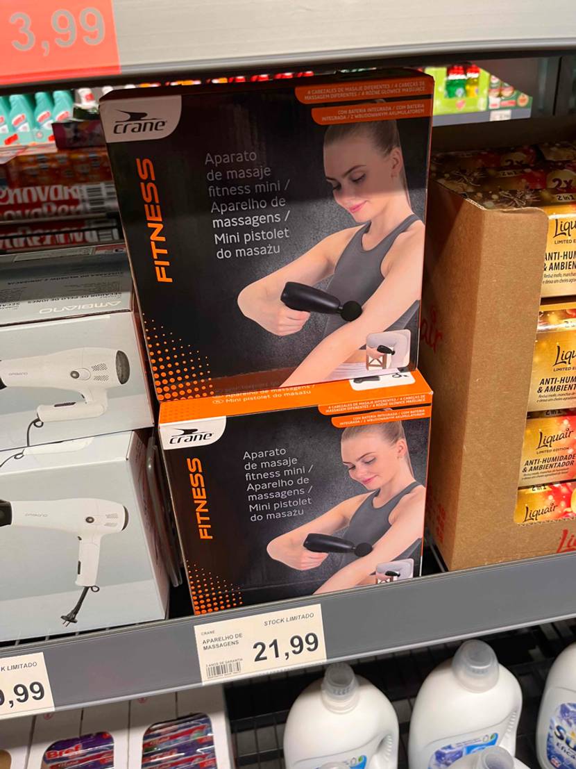 Mehrere originalverpackte Massagepistole im Test stehen in einem Regal mit Preisangabe.