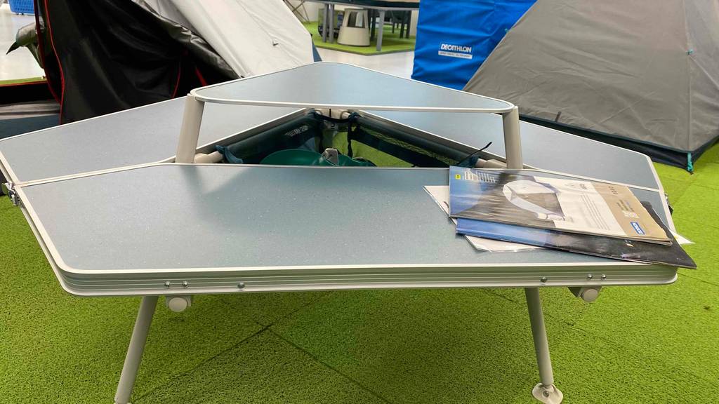 5-Eck-Campingtisch aufgestellt auf einem Rasen mit ausgezogenem Staufach und Prospekten