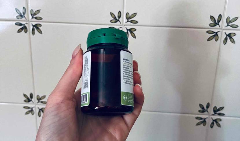 Ginkgo-Tabletten-Test: Hand hält eine lichtdichte Kunststoff-Dose mit grünem Klappdeckel vor einer weißen Fliesenwand mit grünem Blätter-Dekor