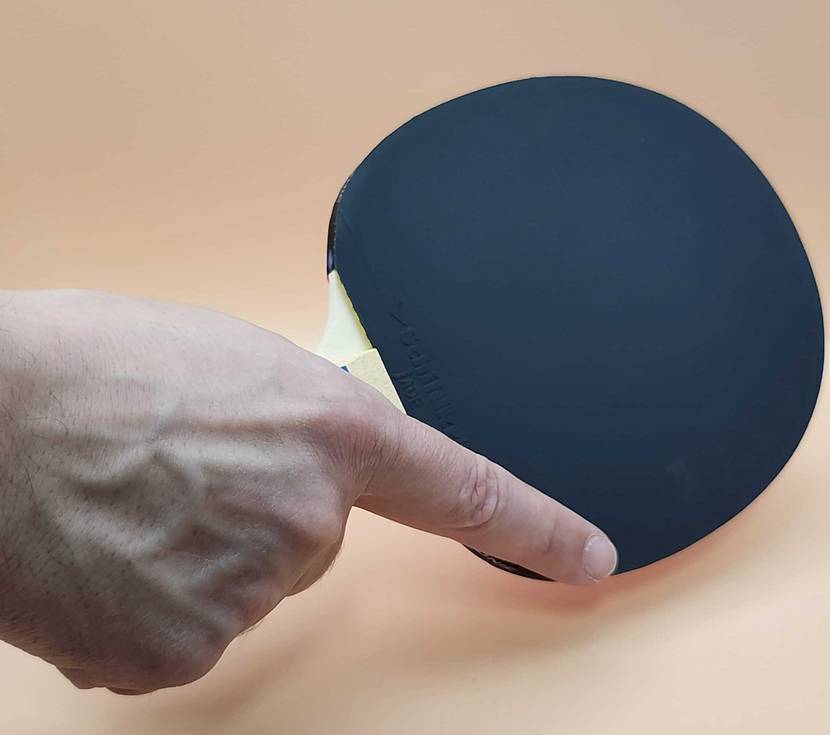Eine Hand hält den getesteten Tischtennisschläger mit der dunklen Beschichtung vor einem beigefarbenen Hintergrund.