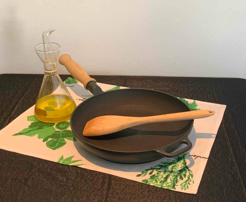 Im Test: Eine Eisenpfanne mit einem Kochlöffel steht neben einer Karaffe Öl auf einer Platzdecke.