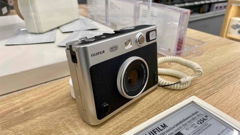 Seitliche Vorderansicht einer schwarz-silbernen Fujifilm Sofortbildkamera im Test, die inklusive Produktschild mit Preisangabe auf einem Verkaufstisch aus Holz ausgestellt ist.