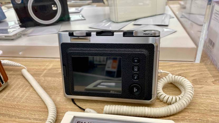 Ansicht des rückwärtigen Bildschirms einer schwarz-silbernen Fujifilm Sofortbildkamera im Test, die gesichert auf einem Ausstellungstisch vor anderen Kameratypen steht.