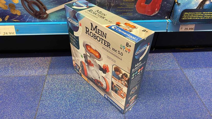 Die Verpackung eines getesteten Roboters für Kinder steht schräg auf einem blauen Boden vor einem Verkaufsregal.