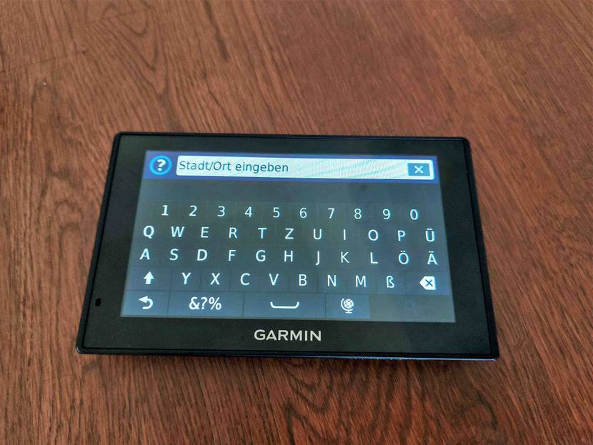 Garmin Navi getestet: ein Gerät liegt eingeschaltet mit Stadteingabeanzeige auf dunklem Boden.