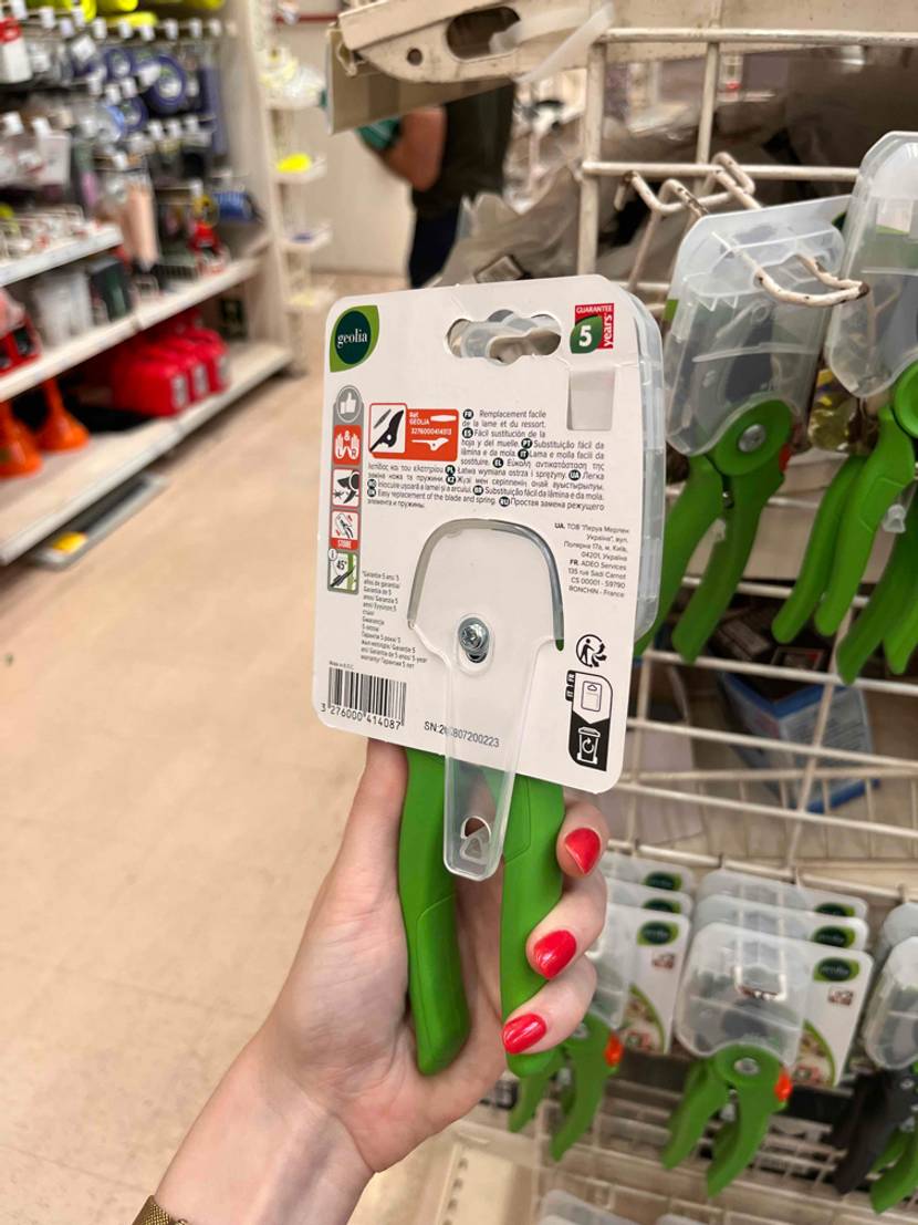 Eine Hand hält eine originalverpackte Gartenschere im Test mit der Rückseite nach vorne.