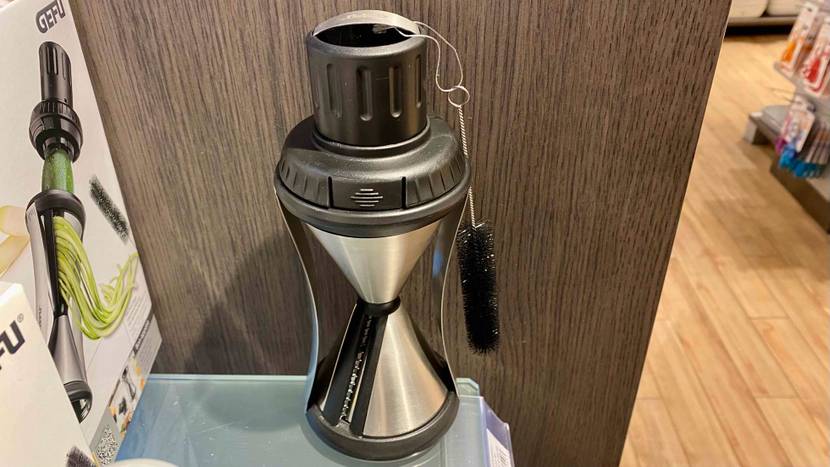 Ansicht von schräg oben auf einen Spiralschneider im Test in schwarz-silberner Metall-Kunststoffausführung neben seiner Originalverpackung auf einem Verkaufsregal.