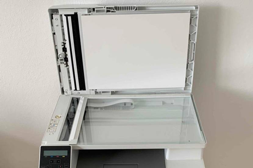 Ein Farblaserdrucker mit geöffnetem Papierfach und offener Scanfläche steht in einem Zimmer auf dem Holzboden.