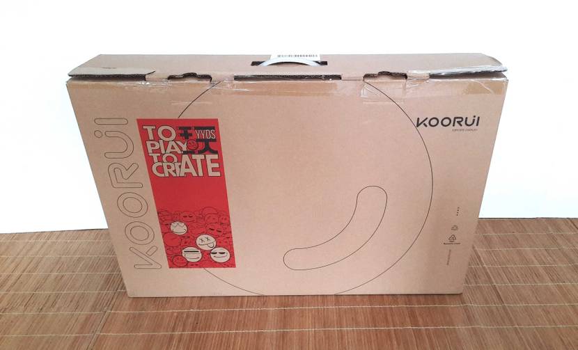 Vorderansicht eines auf einer hellbraunen Bastmatte stehenden, braunen Originalverpackungskartons von einem Koorui 24-Zoll-Monitor im Test.