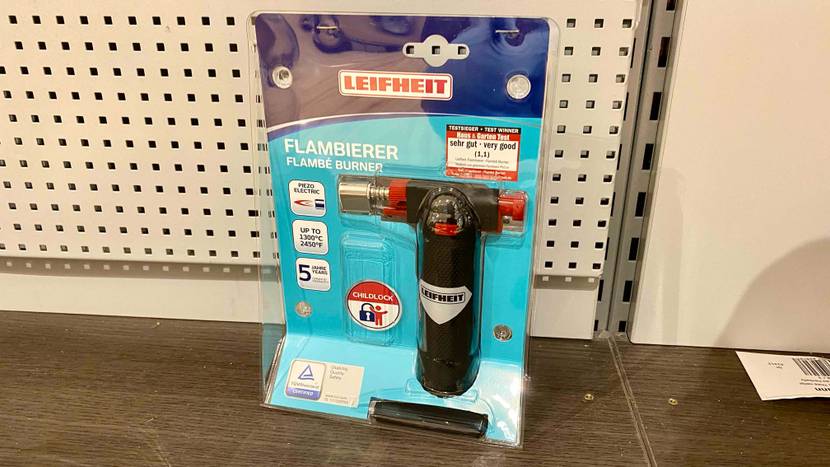 Im Test: Ein originalverpackter Flambierbrenner steht vor einer hellen Metallwand.