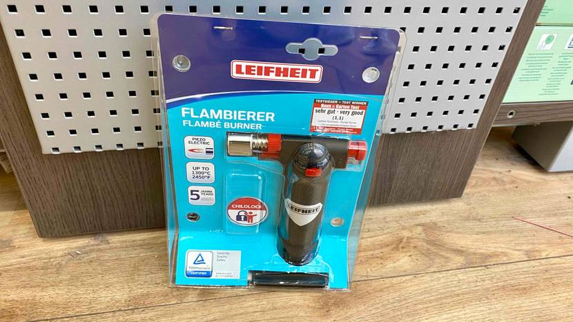 Ein getesteter Flambierbrenner lehnt an einer Fläche aus Holz und Metall auf einem Holzboden stehend.