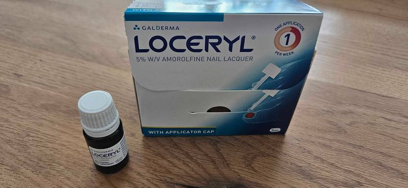 Im Test: Mittel gegen Nagelpilz der Marke Loceryl in einer kleinen, lichtdichten Braunglas-Flasche inklusive weißem Drehverschluss neben der Originalverpackung auf einem Holzuntergrund.