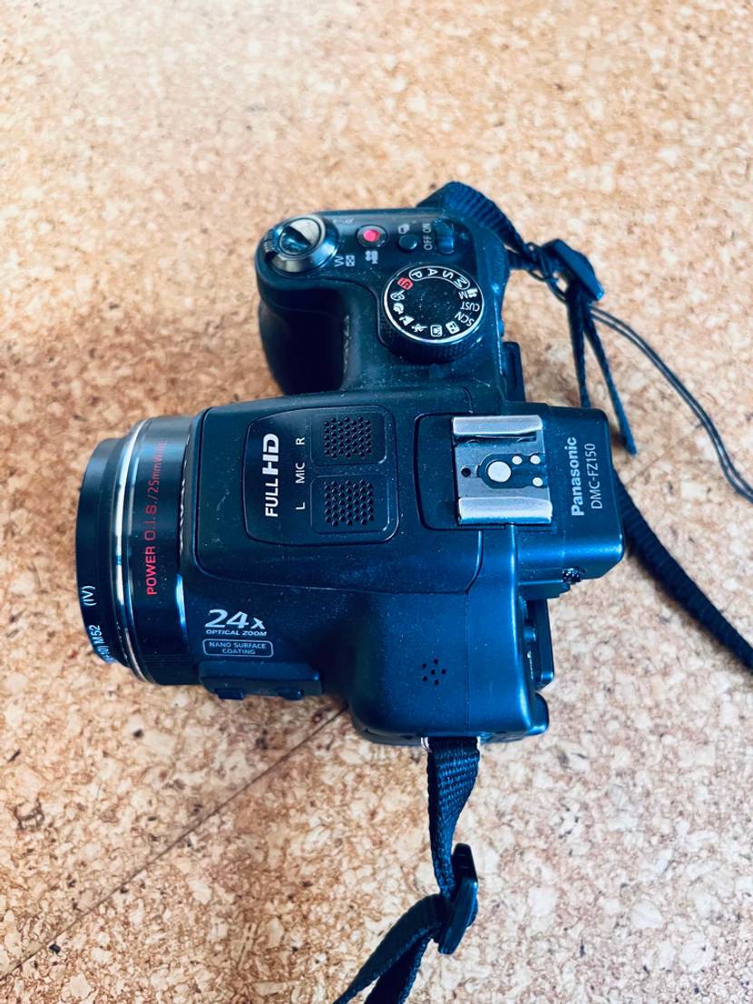 Digitalkamera im Test: Ansicht von oben einer Full-HD Digitalkamera Lumix der Marke Panasonic inklusive Mikrofon sowie Schulterriemen auf einer Korkunterlage.