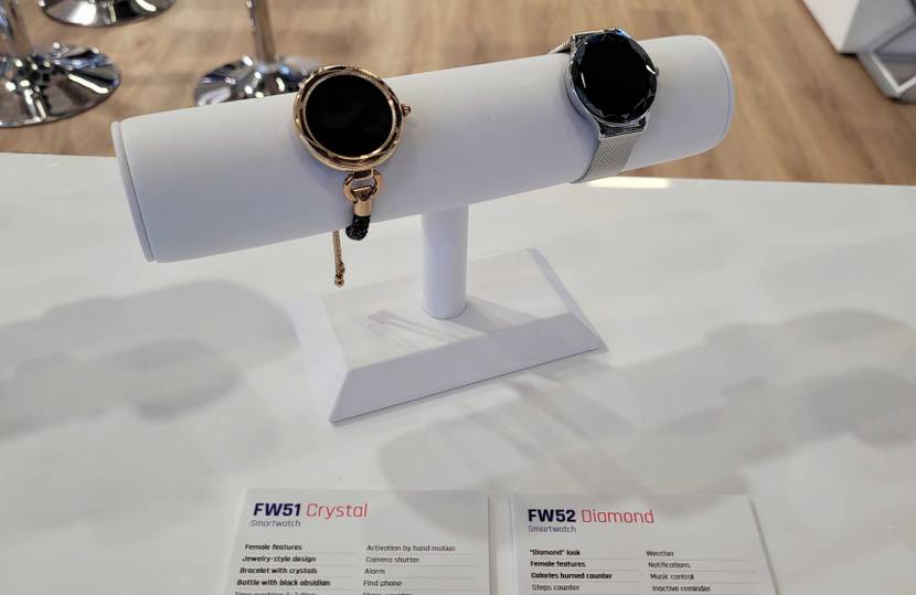 Getestet: Maxcom FW51 Crystal and FW52 Diamond Hybrid Smartwatches auf einem weißen Uhrenhalter mit Beschreibungen im Vordergrund.
