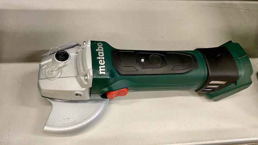 Winkelschleifer im Test: Nahaufnahme von schräg oben auf einen grün-schwarzen Winkelschleifer der Marke Metabo, ohne Akku, inklusive silberfarbener Metall-Schutzhaube auf beigefarbiger Fläche.