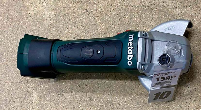 Metabo Winkelschleifer Test: Nahaufnahme von oben auf einen grün-schwarzen Winkelschleifer ohne Akku, mit Schutzhaube aus silberfarbenem Metall und Preisschild auf beigefarbenem Untergrund.