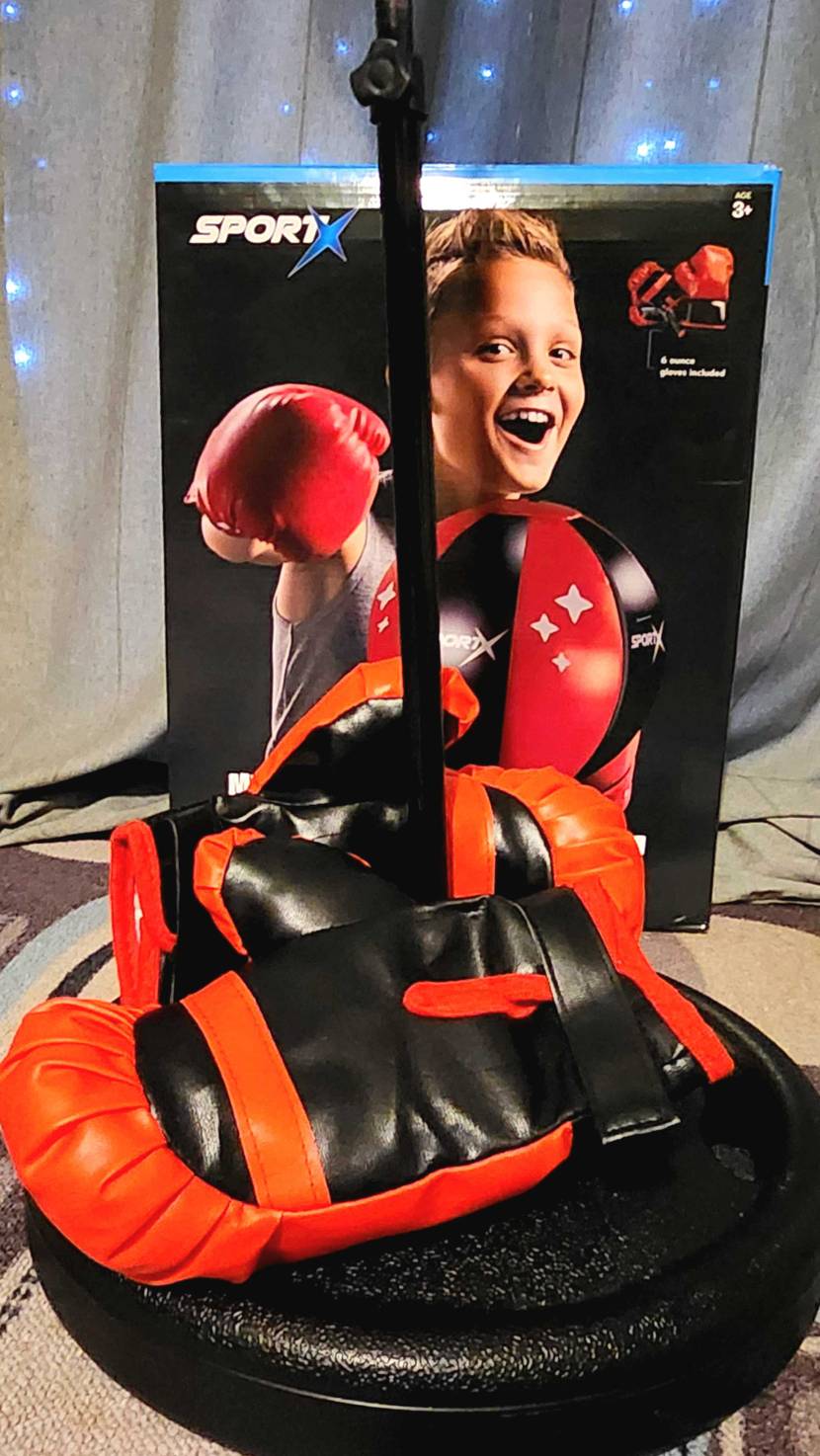 Im Test: Ein Kinder-Boxsack, mit Fokus auf seinen Standfuß, auf dem zwei Boxhandschuhe liegen. Im Hintergrund steht die Boxsack-Verpackung.