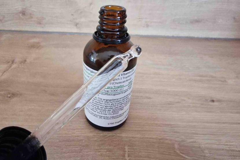 Natural Elements Vitamin-D-Präparat im Test: Geöffnete blickdichte Braunglas-Flasche inklusive Anwendungsbeschreibungs-Etikett und Pipetten-Verschluss vor Holz.