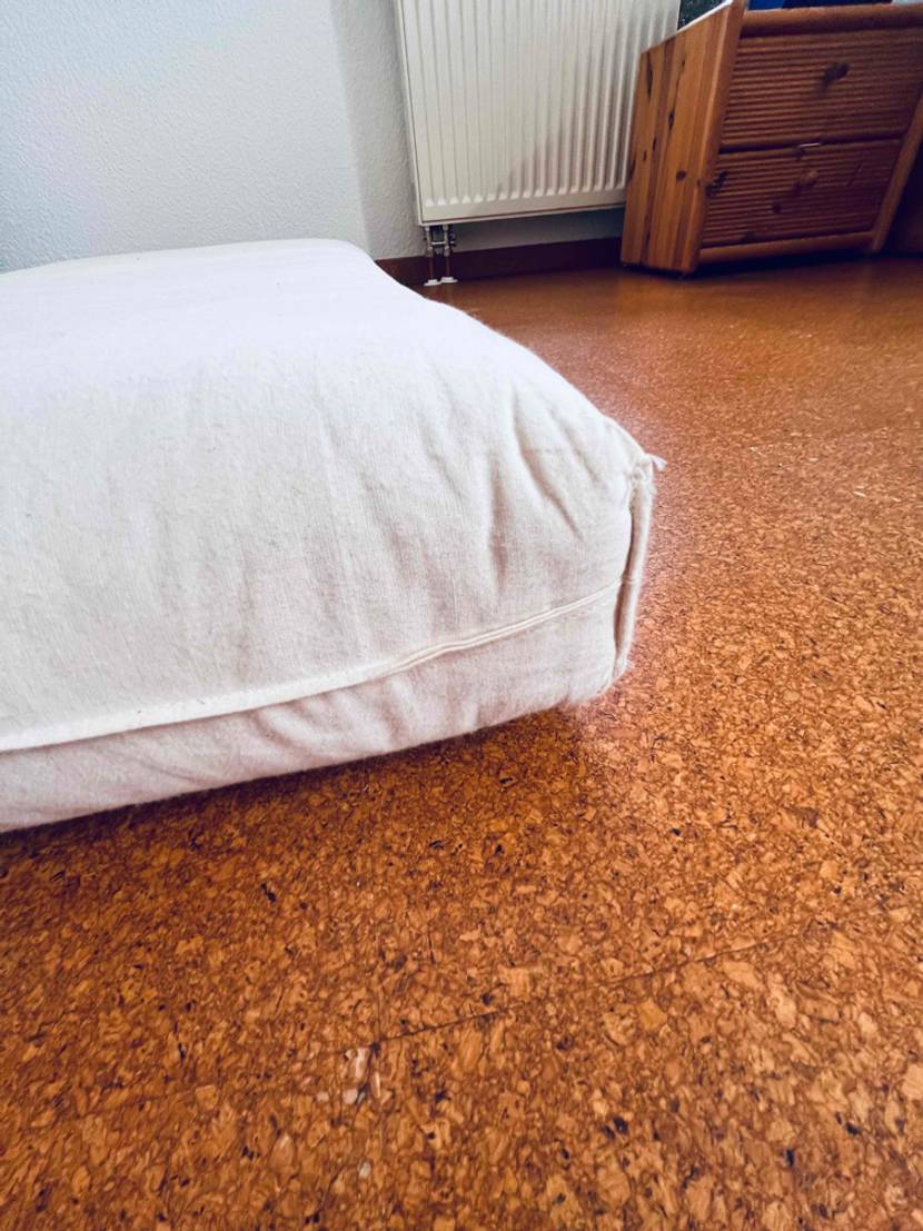 Nahaufnahme einer weißen, getesteten Kindermatratze: Seiten und Ecknähte im Vordergrund auf einem braunen Korkboden mit Holzkommode im Hintergrund.