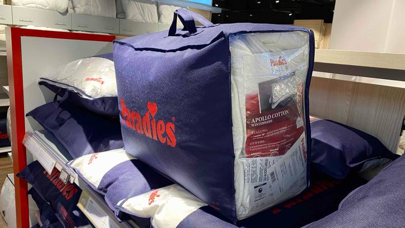 Eine blaue Verpackung inklusive einer Paradies-Thermobettdecke im Test steht inklusive Produktbeschreibung und Zertifikaten auf verpackten Paradies-Kopfkissen in einem Verkaufsbereich