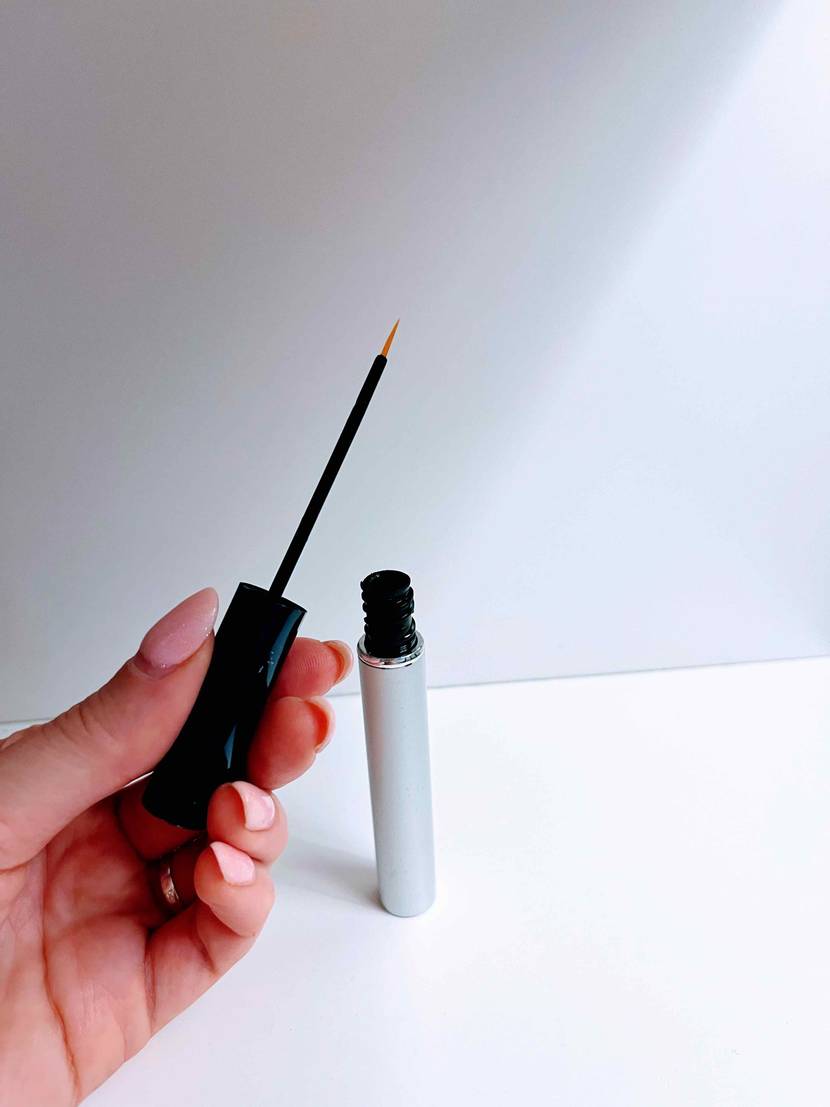 Wimpernserum getestet: Hand hält die Pipette eines geöffneten Fläschchens, das im Hintergrund in weißem Regal steht.