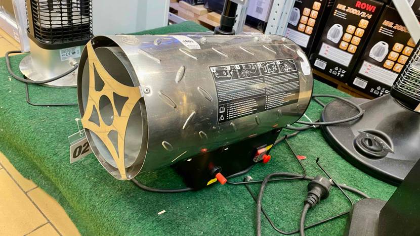Ansicht von der Seite auf einen auf grünem Kunstrasen befindlichen Gasheizstrahler im Test, sein Metall-Zylinder ist auf einer Elektro-Basis inklusive Bedienpanel mit roten Knöpfen befestigt.