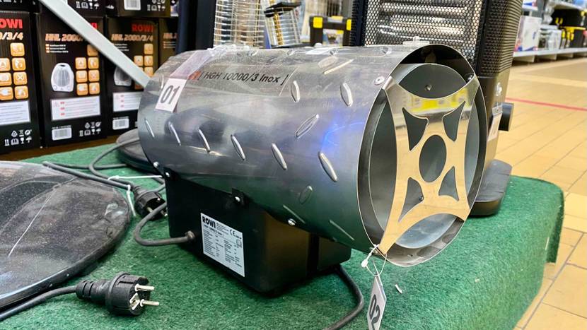 Seitliche Ansicht auf einen auf grünem Kunstrasen im Verkauf ausgestellten ROWI Gasheizstrahler im Test, dessen Metall-Zylinder auf einer schwarzen Elektro-Basis montiert ist.
