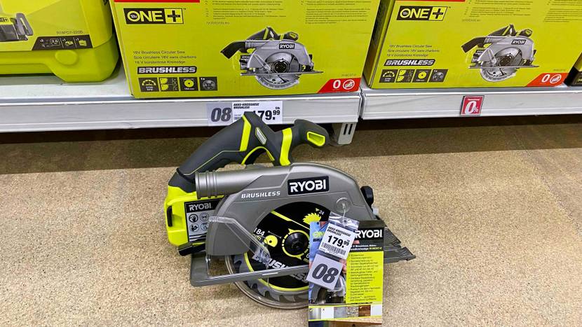Eine bürstenlose gelb-graue Ryobi Kreissäge im Test befindet sich auf einem beigefarbenen Boden unterhalb ihrer Verpackung auf einem Verkaufsregal.
